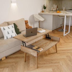 May include: A light wood lift-top coffee table with a laptop and books on it. The table is in a living room setting with a beige sofa, decorative pillows, and a teddy bear. The table is raised, revealing storage space.