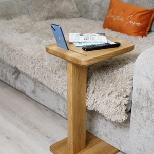 Könnte beinhalten: Ein kleiner, quadratischer Beistelltisch aus Holz mit einem einzelnen, rechteckigen Stützfuß. Ein blaues Smartphone, eine Fernbedienung und eine Zeitschrift liegen auf der Tischoberfläche. Der Tisch steht neben einem grauen Sofa mit einem orangefarbenen Kissen.