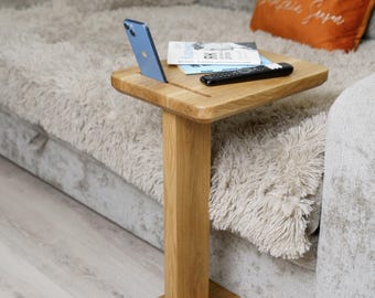 May include: A small, square wooden side table with a single, rectangular support. A blue smartphone, a remote control, and a magazine rest on the table's surface. The table is positioned next to a gray couch with an orange pillow.