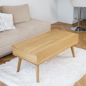 May include: A light oak coffee table with a rectangular top and angled legs, placed on a white fluffy rug. The table's dimensions are approximately 120 cm long. A beige sofa and cushion are in the background.