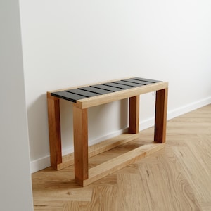 Könnte beinhalten: Moderner Holzbank mit rechteckigem Rahmen und schwarzer Lamellenoberfläche. Die Bank ist aus hellem Holz gefertigt und steht auf einem Fischgrätmusterboden. Das Design ist minimalistisch und modern.