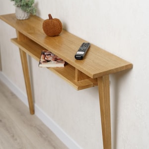 May include: A light oak console table with a lower shelf and angled legs. A small potted plant, a decorative pumpkin, a book, and a remote control are on the table. The table is 48 inches long.