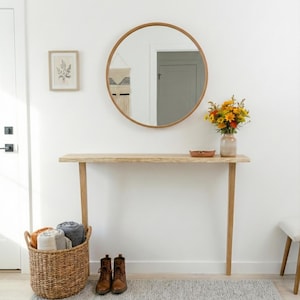 Puede incluir: Una entrada luminosa y espaciosa con un espejo redondo de madera, una consola de madera clara y una cesta de mimbre llena de mantas enrolladas. Un jarrón con flores está sobre la mesa y una alfombra gris está en el suelo. Botas marrones cerca de la alfombra.