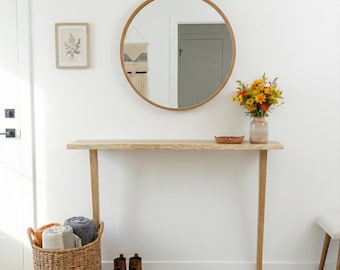 Könnte beinhalten: Ein heller, luftiger Eingangsbereich mit einem runden Holzspiegel, einem Konsolentisch aus hellem Holz und einem Weidenkorb mit gerollten Decken. Auf dem Tisch steht eine Vase mit Blumen, und auf dem Boden liegt ein grauer Teppich. Braune Stiefel stehen in der Nähe des Teppichs.