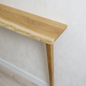 May include: A light-colored wooden console table with a natural edge on one side. The table has a rectangular top and a single, tapered leg. The wood grain is visible. The table is against a white wall.