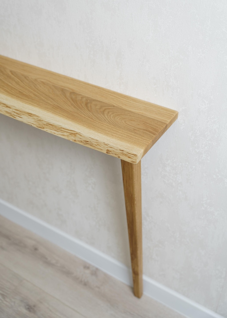 May include: A light-colored wooden console table with a natural edge on one side. The table has a rectangular top and a single, tapered leg. The wood grain is visible. The table is against a white wall.