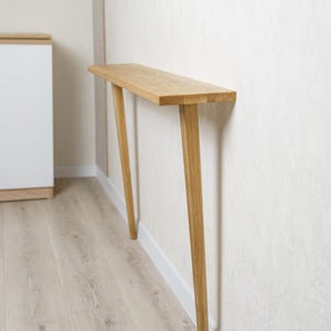 Puede incluir: Una consola de madera clara con una parte superior rectangular y dos patas anguladas. La mesa está colocada contra una pared blanca, mostrando un diseño minimalista. La longitud de la mesa es de aproximadamente 122 cm.