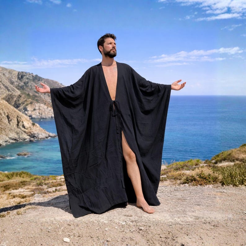 May include: A black, open-front robe with wide sleeves. The robe is knee-length and has a tie closure at the waist. The person is standing on a sandy surface with the ocean and mountains in the background.
