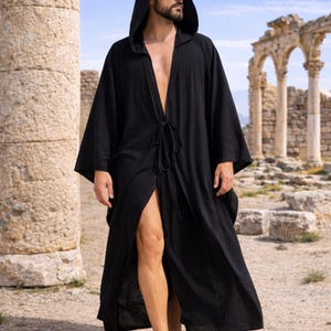 May include: A black hooded robe with a tie closure, worn open, with long sleeves. The robe is made of a flowing fabric. The background features ancient ruins and a blue sky. The robe is suitable for cosplay or casual wear.