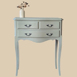 Peut inclure: Une table d'appoint bleu clair à trois tiroirs, avec des pieds courbés et des poignées noires. Un petit vase blanc avec des fleurs séchées est posé dessus. La table a un design classique et vintage.