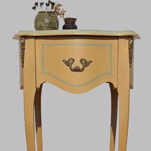 Peut inclure: Petite table d'appoint de style ancien avec un seul tiroir. La table est peinte en beige clair avec un dessus vert menthe et une poignée de tiroir décorative. Des accents dorés ornent les coins. Deux petits vases avec des fleurs séchées sont posés dessus.