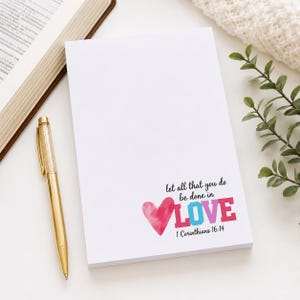 May include: A white notepad featuring a watercolor pink heart and the colorful text "let all that you do be done in LOVE 1 Corinthians 16:14" printed in the bottom right corner, accompanied by a gold metallic pen with a clear crystal top.