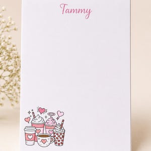 May include: A white rectangular notepad featuring the name Tammy printed in pink at the top. The bottom left corner displays a colorful illustration of various coffee cups and mugs decorated with pink hearts and whipped cream toppings.