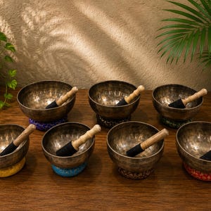 May include: A set of seven metallic singing bowls with hammered textures, each accompanied by a wooden mallet with a black felt tip. The bowls rest on colorful fabric cushions in shades of yellow, blue, red, and green, arranged on a wooden surface.