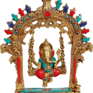 May include: A brass statue of Ganesha sitting on a swing suspended from an ornate arch. The figure and frame are decorated with inlaid red and turquoise stones. The base features a rectangular platform with alternating red and blue stone accents.