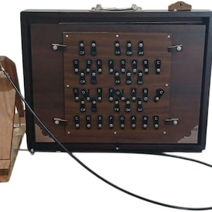 May include: A wooden harmonium with a dark brown finish. The instrument features a keyboard with labeled keys and a foot pedal connected by a black cable. The case has a handle and metal corner accents.