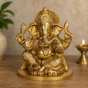 May include: A polished gold-toned brass statue of the deity Ganesha seated in a cross-legged position. The figure features an elephant head, a trunk curled to the left, four arms holding symbolic items, and intricate floral carvings on the clothing.