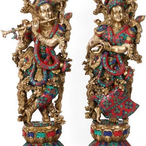 May include: Two ornate brass statues depicting figures in traditional attire, one playing a flute. Both figures feature intricate metalwork inlaid with vibrant red, blue, and green stone accents, standing on decorative lotus-shaped pedestals.