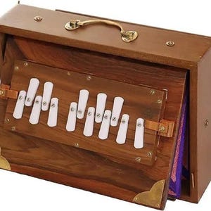 May include: A wooden harmonium with a rectangular shape. The instrument features a keyboard with white keys, a brass handle, and brass corner accents. The wood has a rich, dark brown color.