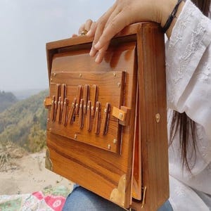 May include: A wooden harmonium with a natural wood finish. The instrument has a rectangular shape with a series of vertical keys and brass corner accents. The harmonium is partially open, revealing the internal components. The background features a scenic view of a mountain range.