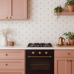 Puede incluir: Una cocina moderna con horno negro, encimera de gas y armarios rosas. Una encimera blanca sostiene una jarra blanca, una taza marrón y una tetera dorada. Plantas en macetas sobre una estantería y encimera, con papel pintado floral.