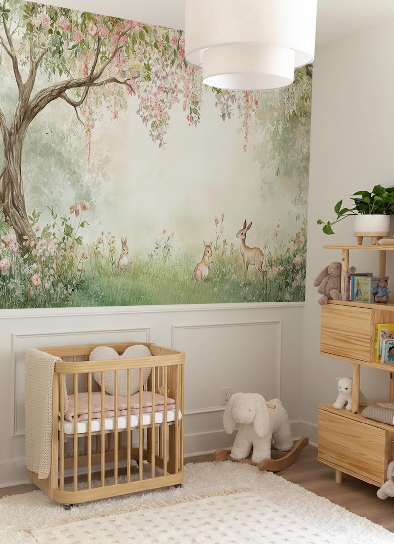 May include: A nursery featuring a light wood crib, a white rug, and a wall mural of a forest scene with deer and rabbits. A wooden bookshelf holds books and soft toys. A white rocking bunny sits on the floor.