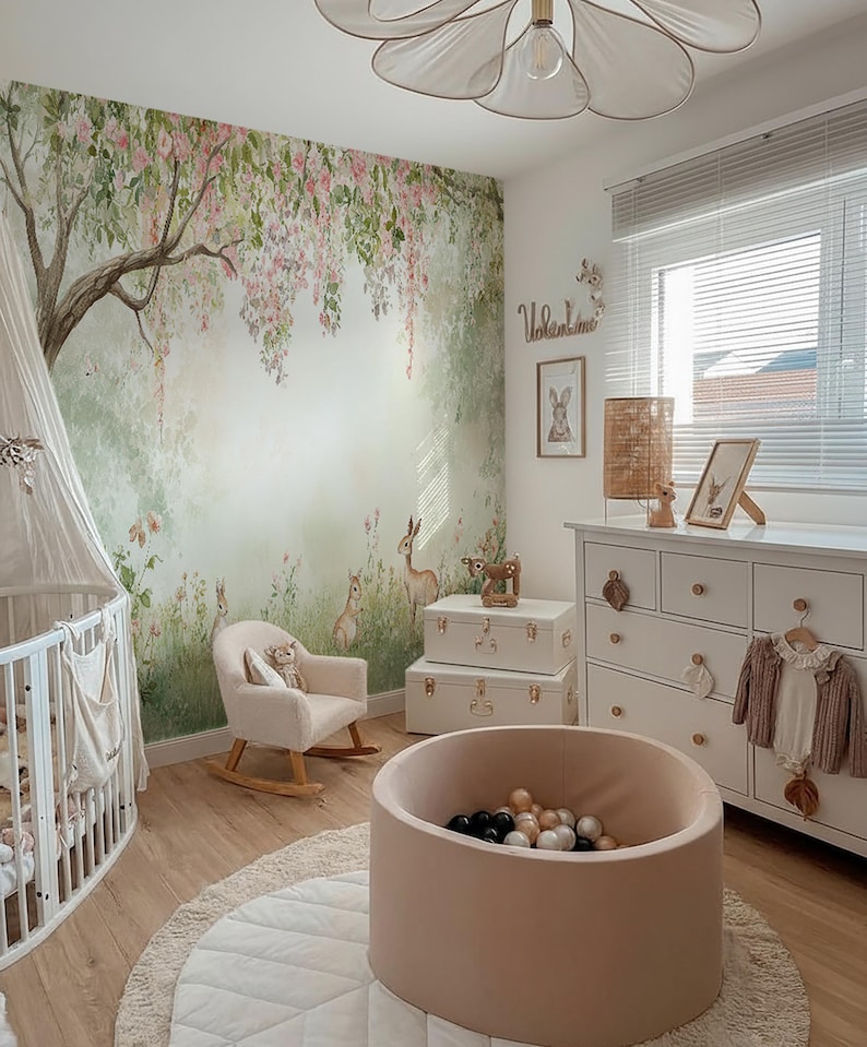 May include: A child's nursery featuring a forest mural, a white cot, and a pink rocking chair. A ball pit with black and white balls is on a round rug. A white chest of drawers and storage trunks complete the room's decor.
