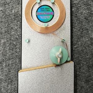 Puede incluir: Una herramienta Table Foiler para el laminado de vidrio. La herramienta tiene un rollo de cinta de cobre, una bobina de plástico verde y una etiqueta que dice "Foils Glass - First Class". La herramienta está montada en una tabla rectangular.