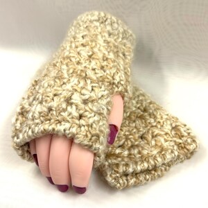 May include: A pair of beige and cream-colored crocheted fingerless gloves. The gloves are made with a thick, textured yarn and have a hole for the thumb. The gloves are designed to keep hands warm while leaving fingers free.