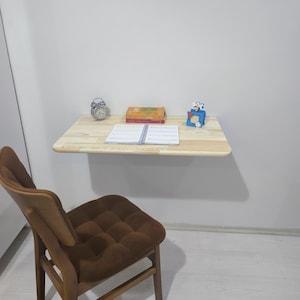 May include: A light-colored wooden wall-mounted desk with a small alarm clock, books, and a notebook. A brown chair with a tufted seat is in front of the desk. A small blue decorative item is on the desk.