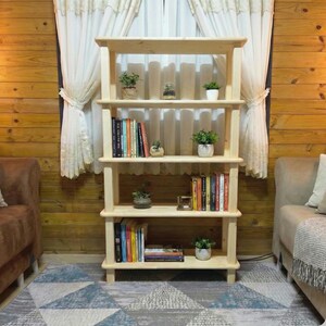 May include: A light-colored wooden bookshelf with four shelves. The shelves hold books and small potted plants. The bookshelf is against a wood-paneled wall, with a patterned rug on the floor. Curtains are visible in the background.
