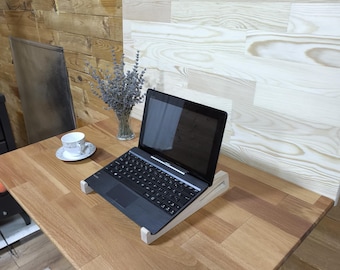 May include: A wooden desk with a laptop on a wooden stand, a teacup, and a vase of lavender. The desk is made of light brown wood and has a rounded edge. The laptop is black and open, and the stand is made of light-colored wood.