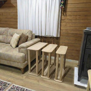 May include: Three light-colored wooden side tables with rectangular tops and four supporting legs each. The tables are arranged in a row, with a beige sofa and pillows in the background. The room has wood paneling and a white curtain.
