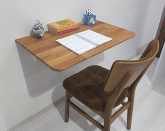 May include: A wall-mounted wooden desk with a light brown finish, featuring a silver analog alarm clock, a stack of two books, a blue pen holder, and an open spiral-bound planner. A wooden chair with a brown tufted cushion sits in front.
