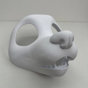 May include: A white, unpainted animal mask base featuring large eye openings, a prominent snout, and a slightly open mouth revealing a row of small, molded teeth. The surface has a smooth, matte finish with visible 3D printed texture lines.