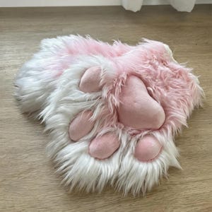 May include: A pair of plush animal paw gloves featuring long, soft white and light pink faux fur. The palm side displays four rounded, smooth, light pink fabric paw pads and a larger central pad, all stitched into the furry material.