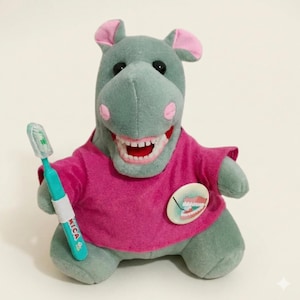 May include: A plush hippo toy in shades of gray and pink, wearing a pink shirt and holding a teal toothbrush. The hippo has a large, open mouth with visible teeth. A round badge with a tooth graphic is on the shirt.