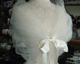 May include: A white tulle bridal shawl with a satin ribbon tied in a bow at the back. The shawl is draped over a white dress form.