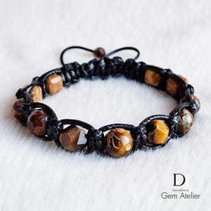 Könnte beinhalten: Handgefertigtes Makramee-Armband mit braunen und beigefarbenen Edelsteinperlen. Das Armband ist mit schwarzem Kordelmaterial gefertigt und hat einen Schiebeknotenverschluss. Die Perlen umfassen Tigerauge und andere Natursteine. Gem Atelier Logo.