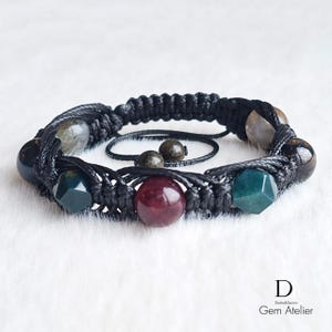 May include: A black macrame bracelet with a variety of gemstone beads in shades of green, red, and gray. The bracelet features a central design with a smaller, matching bracelet and two additional beads. The Gem Atelier logo is visible in the bottom right corner.
