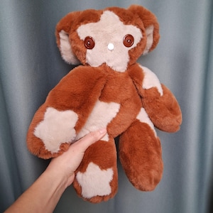 Può includere: Un orsacchiotto di peluche fatto a mano con pelliccia marrone e crema. L'orso ha occhi a bottone e un naso bianco. Toppe a forma di stella color crema adornano le zampe e il petto. L'orso è tenuto su uno sfondo blu.
