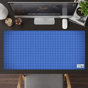 May include: Blue desk mat with a white grid pattern, resembling a blueprint. The mat has the word "HIT" in white in the bottom right corner. The mat is rectangular and covers a large portion of the desk.