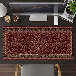 May include: A rectangular desk mat with a traditional rug design. The mat features a deep red background with intricate floral and geometric patterns in shades of beige, gold, and black. The mat is placed on a dark desk, with a computer monitor, mouse, and drinks visible.