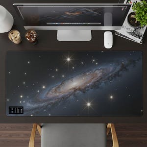 May include: A rectangular desk mat featuring a detailed image of a galaxy with swirling blues, browns, and white hues. The mat has a black border and the word "HIT" in a black box in the lower left corner. The mat is on a dark brown desk.