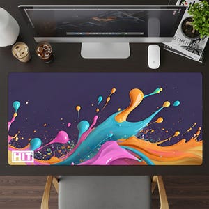 May include: A rectangular desk mat with a vibrant abstract design. The mat features a dark purple background with colorful splashes of pink, orange, and turquoise paint. The word "HIT" is in a white box in the lower left corner.