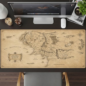 May include: A rectangular desk mat featuring a vintage-style map of Middle-earth printed in black ink on a tan, parchment-colored background. The map displays detailed geographical features, mountain ranges, and labels including Mordor and Gondor.