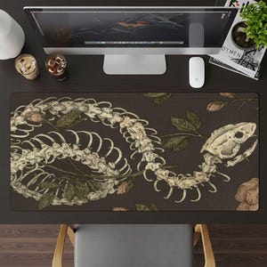 May include: A dark brown desk mat with a detailed illustration of a snake skeleton entwined with leaves and flowers. The design uses cream, brown, and green, creating a vintage aesthetic. The mat is rectangular and designed to protect a desk surface.