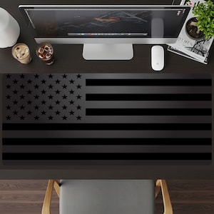 May include: A rectangular desk mat featuring a monochromatic black and dark gray American flag design. The mat displays a field of stars on the left side and horizontal stripes extending across the remainder of the surface in a subtle, low-contrast style.