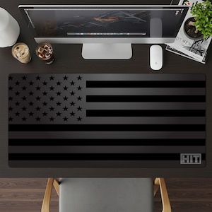 May include: Black desk mat featuring a stylized American flag design. The mat has a matte finish and a rectangular shape. The flag design includes stars and stripes in shades of black. The word "HIT" is in the lower right corner.