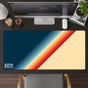 May include: A rectangular desk mat with a retro design. The mat features a dark blue section on the left and a cream-colored section on the right, separated by diagonal stripes in shades of blue, orange, and red. The word "HIT" is in a white box in the lower left corner.